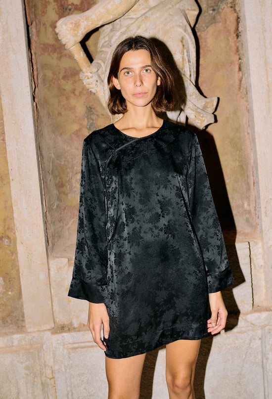 Cropped knees up of woman wearing a black floral patterned long sleeve mini dress standing in an architectural setting.