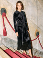 Woman wearing a black velvet dress with long sleeves and front waist tie / tassels standing against a marble wall with red accents.