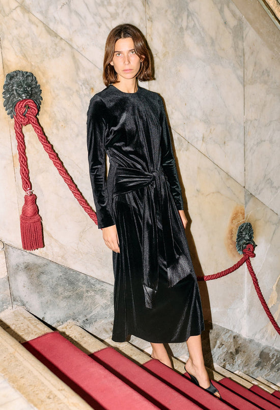 Woman wearing a black velvet dress with long sleeves and front waist tie / tassels standing against a marble wall with red accents.