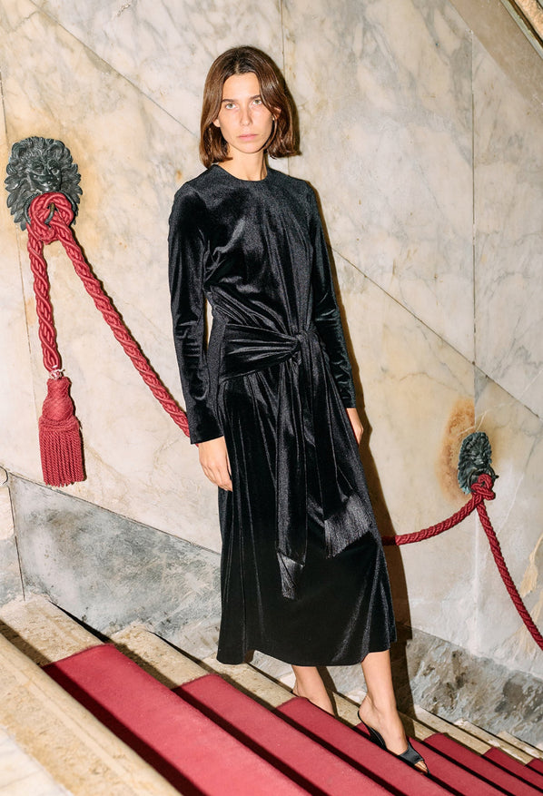 Woman wearing a black velvet dress with long sleeves and front waist tie / tassels standing against a marble wall with red accents.