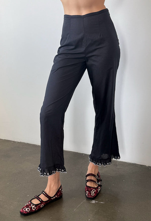 Cropped waist down of woman wearing black pants with ruffled hem and floral shoes on a plain background