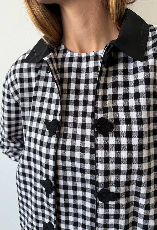 Cropped chin to stomach image of woman wearing a black and white checkered gingham jacket with black embroidered buttons and collar with matching under dress on a plain background