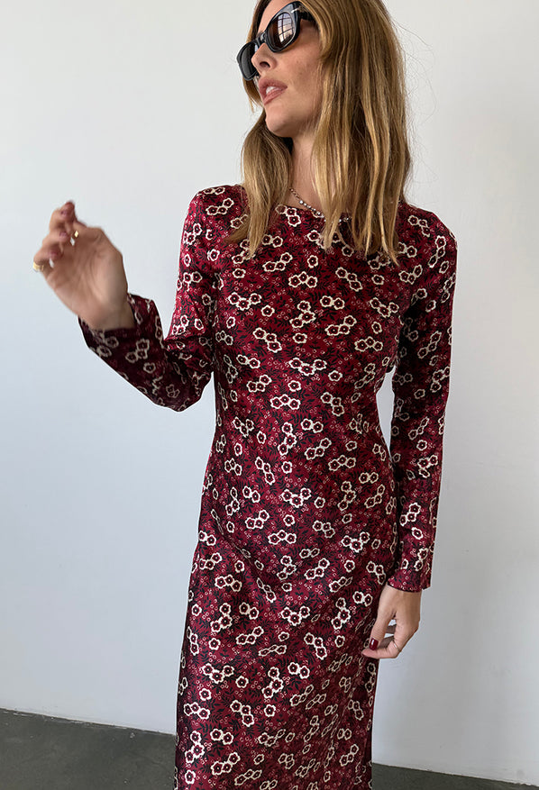 Cropped image of woman wearing a silky midi length long sleeve ruby floral print dress against a plain background