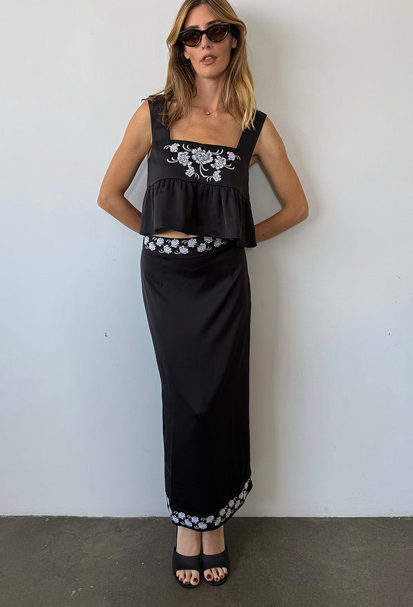 Woman wearing a black sleeveless top with floral embroidery and loose ruffled waist and a matching black satin skirt with floral embroidery on waist and hem against a white wall.