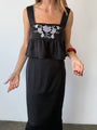 Image cropped neck to knee of woman wearing a black sleeveless top with floral embroidery and loose ruffled waist and a matching black satin skirt against a white wall.