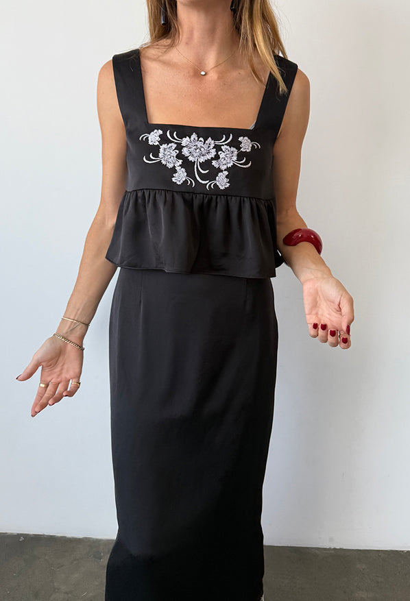 Image cropped neck to knee of woman wearing a black sleeveless top with floral embroidery and loose ruffled waist and a matching black satin skirt against a white wall.