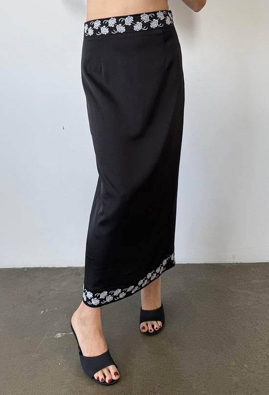 Cropped waist down of woman wearing a black satin ankle length skirt with white embroidered flowers on the waist and hem and black shoes against a white background