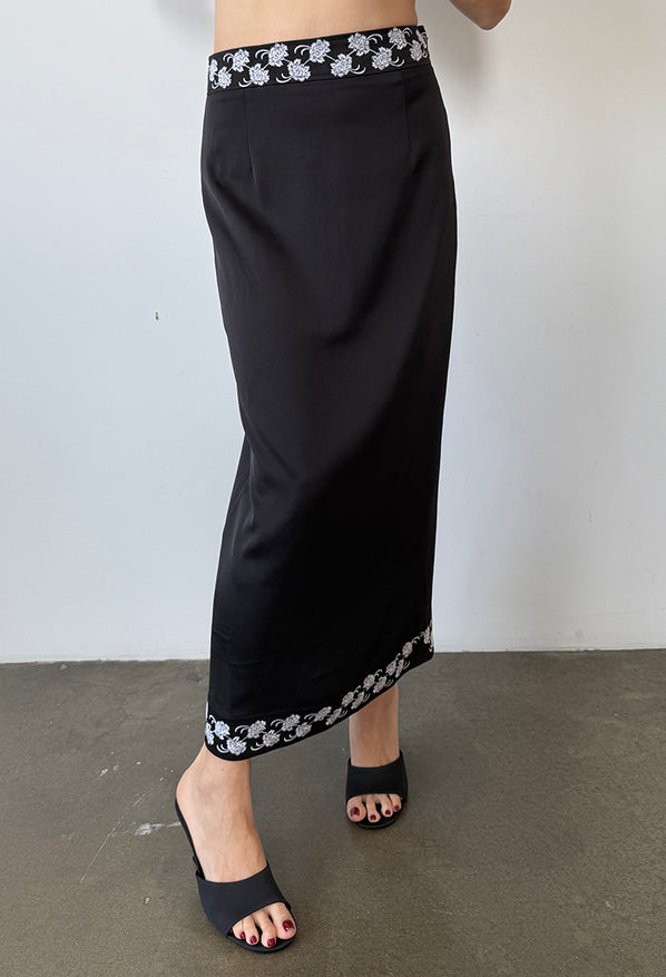 Cropped waist down of woman wearing a black satin ankle length skirt with white embroidered flowers on the waist and hem and black shoes against a white background