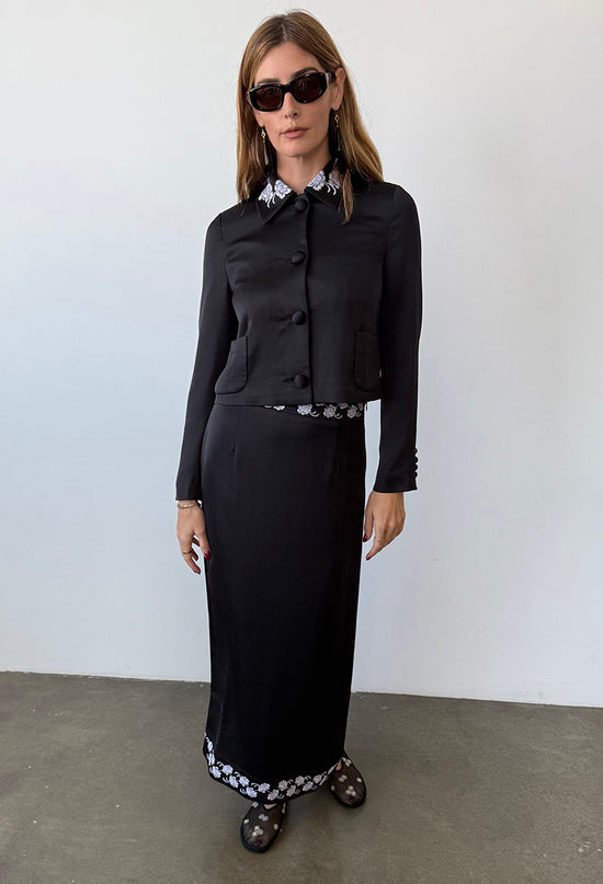 Woman wearing a black satin jacket with white flowers embroidered on the collar with a matching black ankle length skirt with white embroidered flowers on the waist and hem against a white background