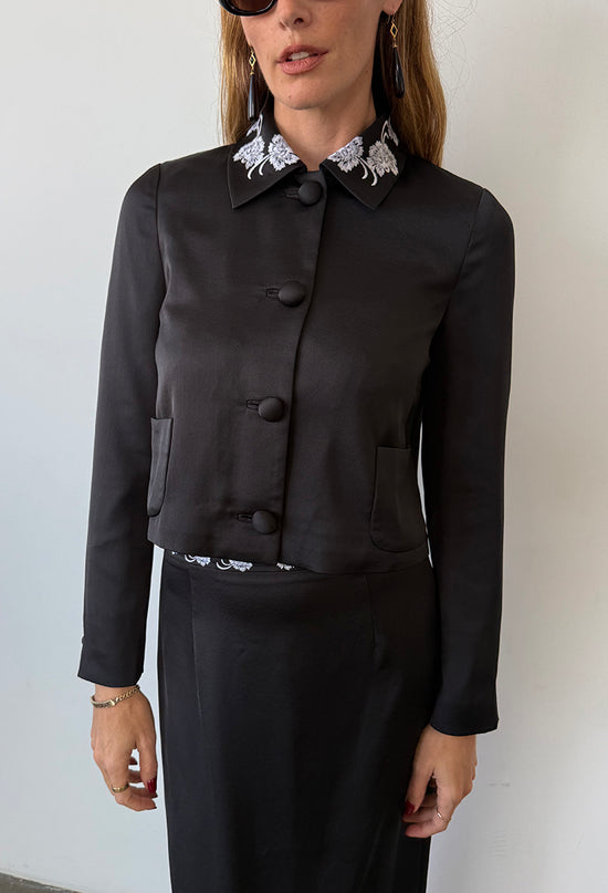 Cropped nose to thigh image of woman wearing a black satin jacket with white flowers embroidered on the collar with a matching black skirt with white embroidered flowers on the waist against a white background