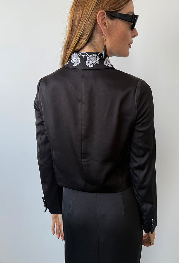 Cropped image of woman wearing a black satin jacket with white flowers embroidered on the collar with a matching black satin skirt against a white background