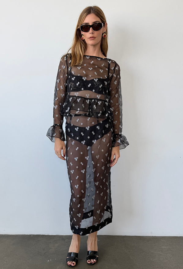 Woman wearing a black sheer long sleeve top with small embroidered flowers, ruffle around wrist and hem paired with matching sheer black skirt with small embroidered flowers against a plain background. 