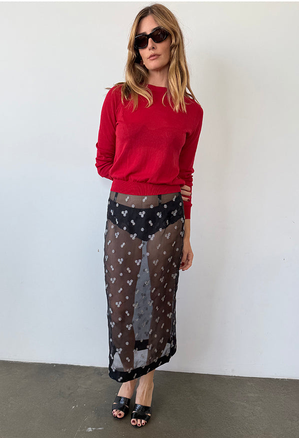 Woman wearing a red sheer knit pullover sweater and sheer black skirt patterned with small embroidered flowers against a white wall