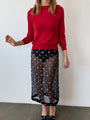 Woman wearing a red sheer knit pullover sweater and sheer black skirt patterned with small embroidered flowers against a white wall
