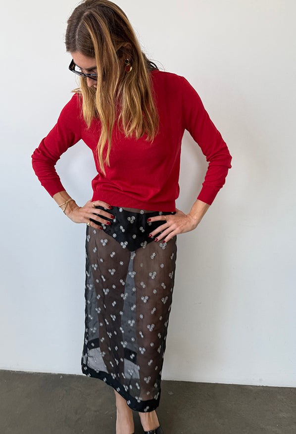 Slightly cropped image of woman wearing a red sheer knit pullover sweater and sheer black skirt patterned with small embroidered flowers against a white wall