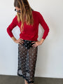 Woman wearing a red sheer knit pullover sweater and sheer black skirt patterned with small embroidered flowers against a white wall