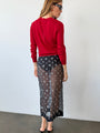 Behind view of woman wearing a red sheer knit pullover sweater and sheer black skirt patterned with small embroidered flowers against a white wall