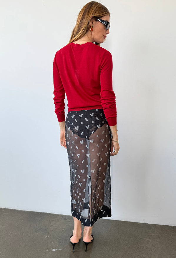 Behind view of woman wearing a red sheer knit pullover sweater and sheer black skirt patterned with small embroidered flowers against a white wall