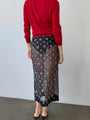 Cropped from the neck down, back side view of woman wearing a red sheer knit pullover sweater and sheer black skirt patterned with small embroidered flowers against a white wall