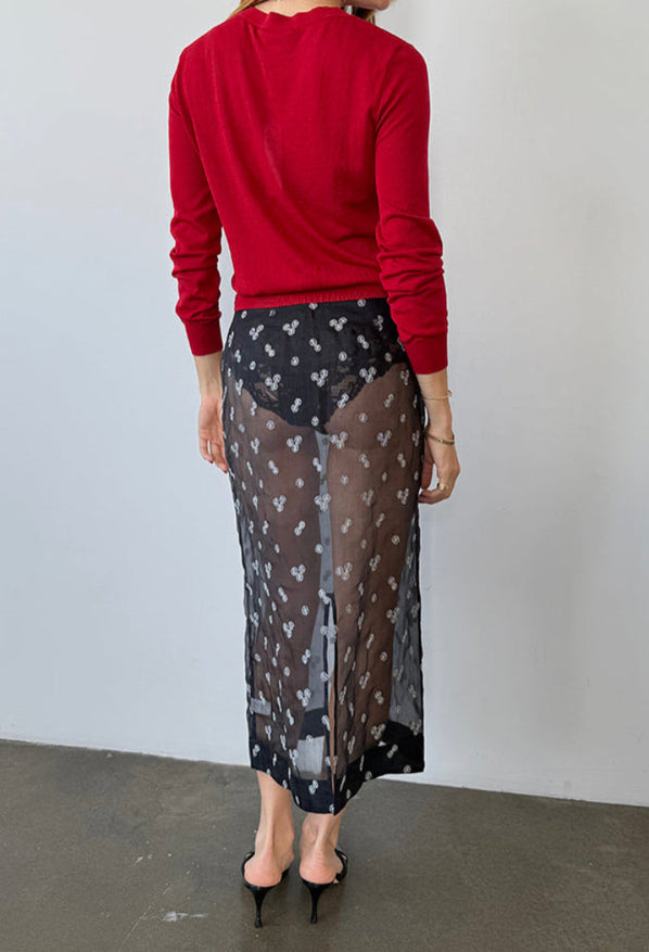 Cropped from the neck down, back side view of woman wearing a red sheer knit pullover sweater and sheer black skirt patterned with small embroidered flowers against a white wall