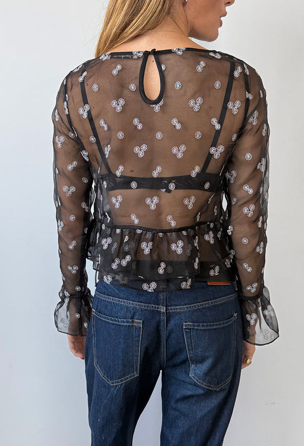 Back view cropped nose to thigh of woman wearing a black sheer long sleeve top with small embroidered flowers and ruffles around wrist and hem with blue jeans against a plain background