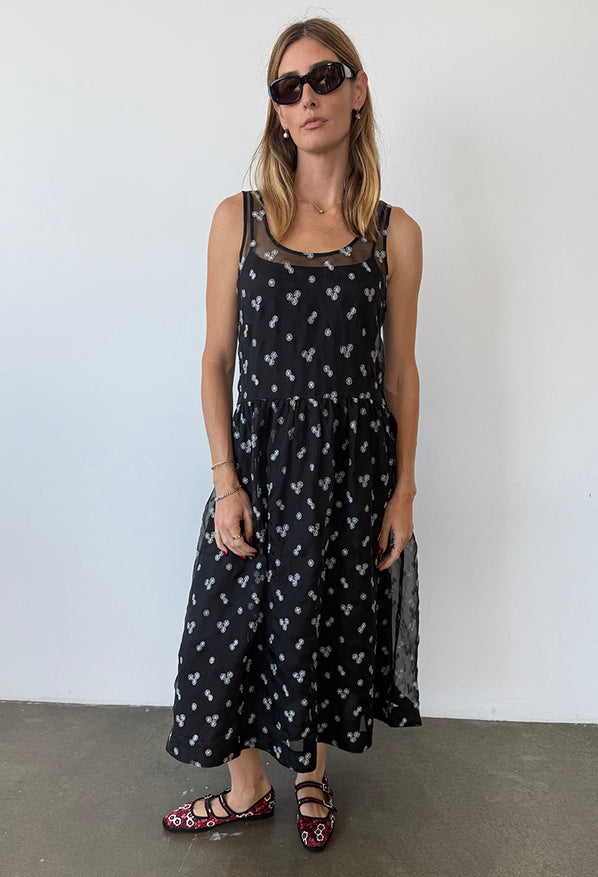 Woman wearing a long black sleeveless sheer dress with small white floral embroidery and drop waist against a plain background