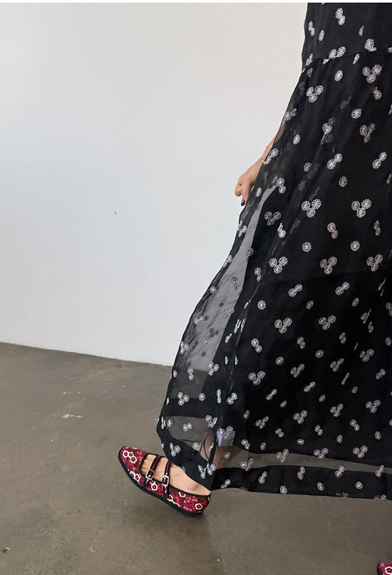 Cropped waist down of woman walking wearing a long black sheer dress with small white floral embroidery and drop waist against a plain background