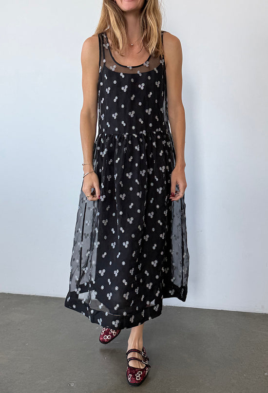 Cropped chin down of woman wearing a long black sleeveless sheer dress with small white floral embroidery and drop waist against a plain background