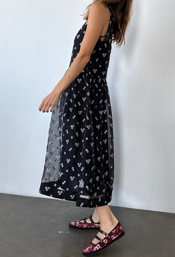 Cropped shoulder down side view of woman wearing a long black sleeveless sheer dress with small white floral embroidery and drop waist against a plain background