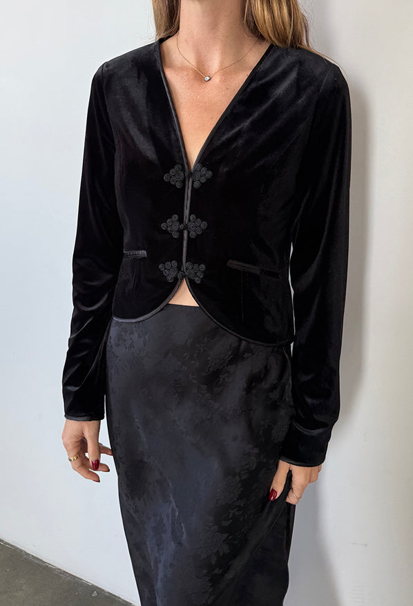 Image cropped neck to thigh of woman wearing a black velvet jacket / top and black floral silky skirt against a white wall.