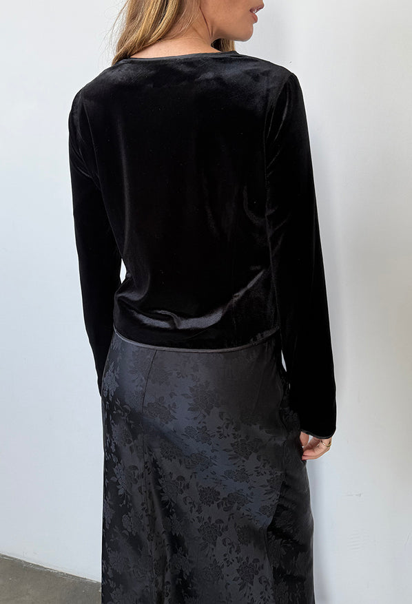 Cropped image nose to thigh behind view of woman wearing a black velvet jacket / top and black floral silky skirt against a white wall.