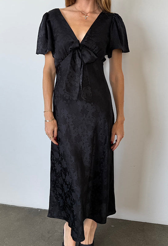 Woman in a long black floral print dress with ruffled short sleeves and a front bow detail against a plain background