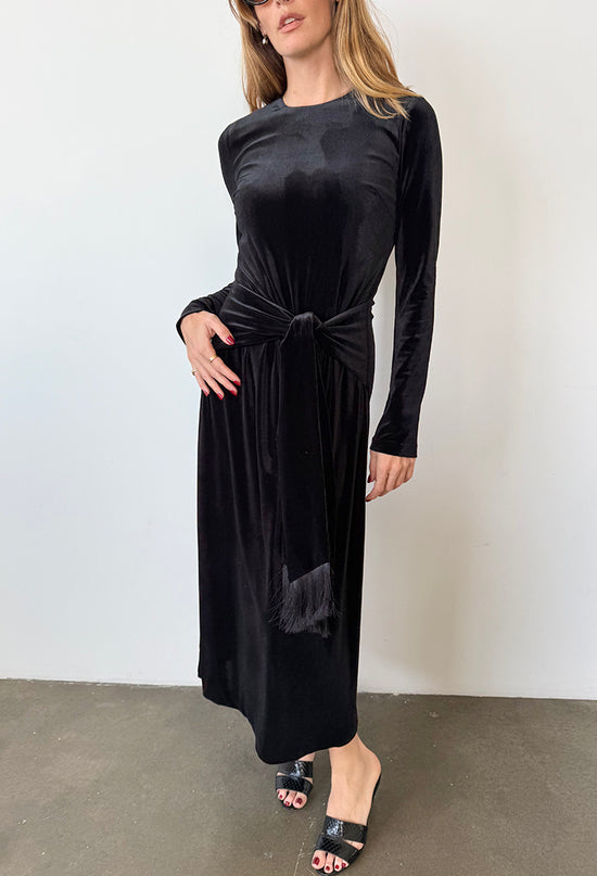 Woman wearing a black velvet dress with long sleeves and front waist tie / tassels against a white background