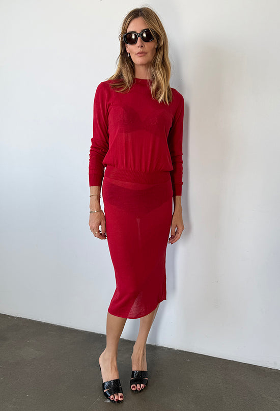 Woman wearing a red pullover sheer knit cardigan with a red sheer midi skirt against a white wall