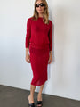 Woman wearing a red sheer knit pullover sweater and sheer red knit skirt against a white wall