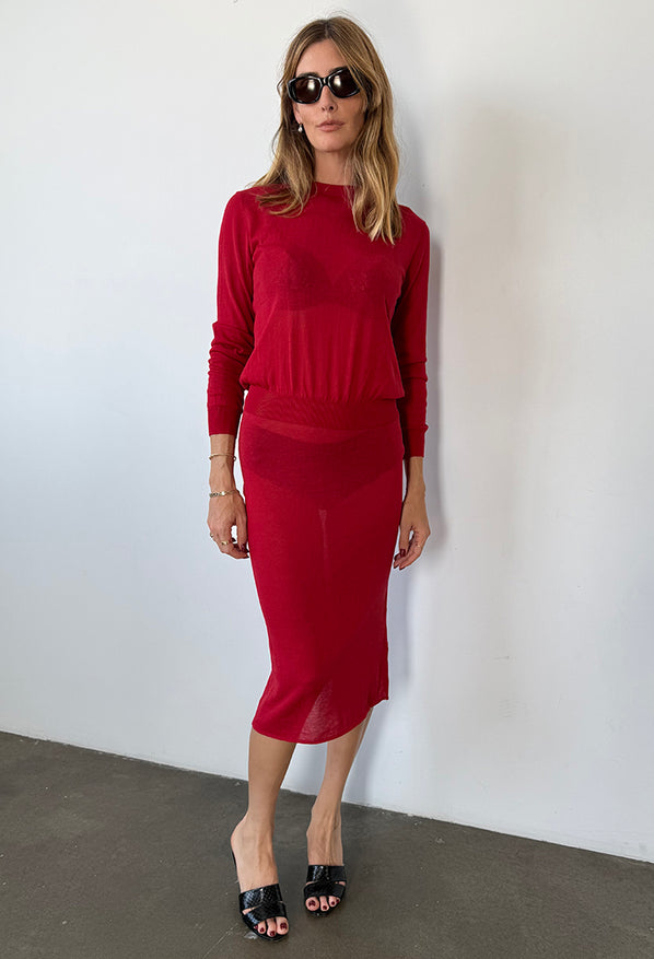 Woman wearing a red sheer knit pullover sweater and sheer red knit skirt against a white wall