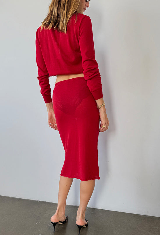 Backside view of woman wearing a red pullover sheer sweater with a red sheer midi skirt against a white wall