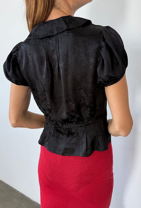 Cropped chin to thigh, back view of woman wearing a black floral patterned blouse with short sleeves and red sheer knit skirt against a white background