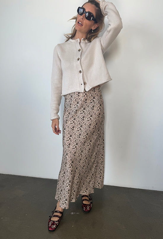 Woman wearing a beige button up cardigan, silk and viscose skirt with floral cherry blossom pattern, and black sunglasses against a plain background
