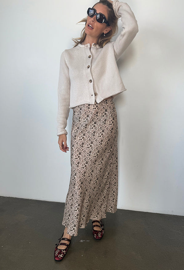 Woman wearing a beige button up cardigan, silk and viscose skirt with floral cherry blossom pattern, and black sunglasses against a plain background