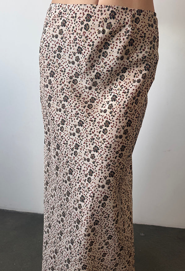 Cropped waist down of woman wearing a long silk and viscose skirt with floral cherry blossom pattern, and red floral shoes against a plain background