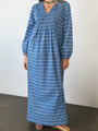 Cropped neck down of a woman in a long sleeve blue Caftan dress with a v-neck and zigzag print standing against a plain background.