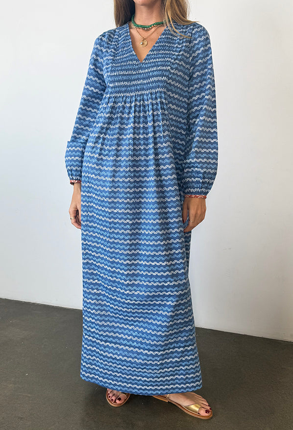 Cropped neck down of a woman in a long sleeve blue Caftan dress with a v-neck and zigzag print standing against a plain background.