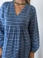 Cropped neck to waist of a woman in a long sleeve blue Caftan dress with a v-neck and zigzag print standing against a plain background.