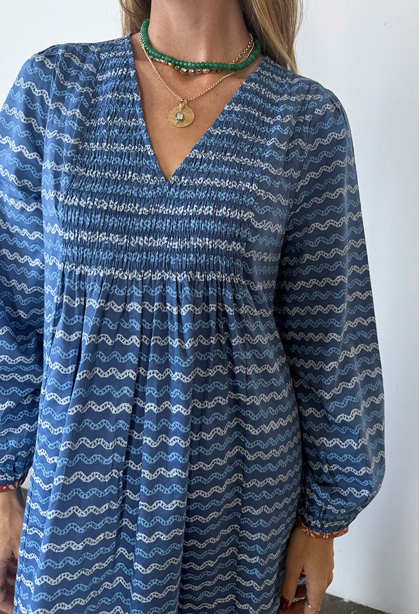 Cropped neck to waist of a woman in a long sleeve blue Caftan dress with a v-neck and zigzag print standing against a plain background.
