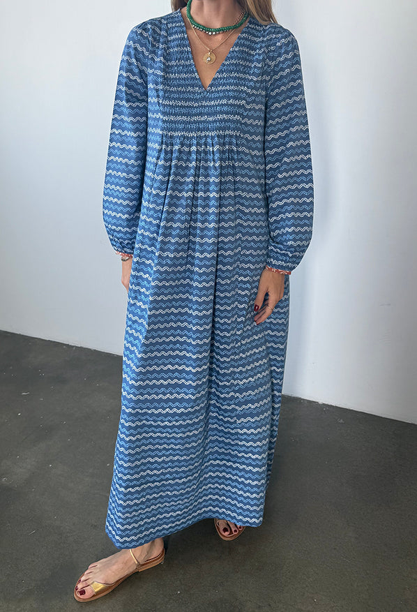 Cropped neck down of a woman in a long sleeve blue Caftan dress with a v-neck and zigzag print standing against a plain background.