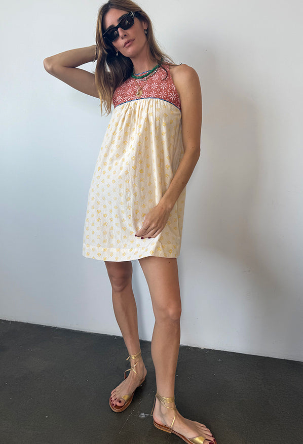 Woman wearing a mini length tank style shift dress with blood orange and white floral top and white skirt with yellow sun pattern standing in front of plain background. 