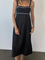 Cropped neck down of woman in a black midi length tank dress with lace bodice and white embroidered ruffle trim details standing against plain background. 
