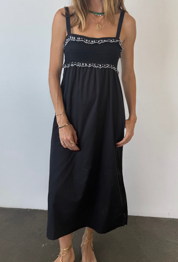 Cropped neck down of woman in a black midi length tank dress with lace bodice and white embroidered ruffle trim details standing against plain background. 