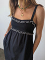 Cropped neck to waist of woman in a black midi length tank dress with lace bodice and white embroidered ruffle trim details standing against plain background. 